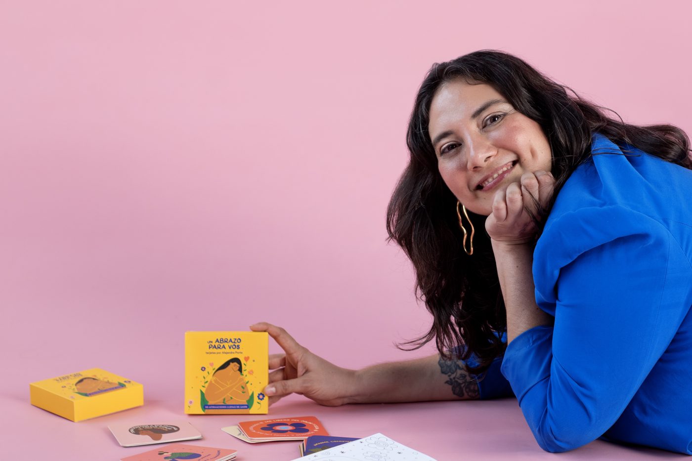 Ale recostada en un piso rosa con sus cartas de afirmación y su libro para pintar